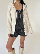 A model wearing a cream-colored, fuzzy knit cardigan over a floral tank and short set, paired with cream-colored knee-high boots, against a plain off-white background.