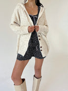 A model wearing a cream-colored, textured knit cardigan over a set, paired with beige boots, against a plain off-white background.