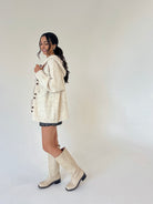 A woman modeling a cream-colored knit cardigan with brown buttons, worn over a dark patterned dress and paired with matching cream knee-high boots, set against a plain light gray studio background.