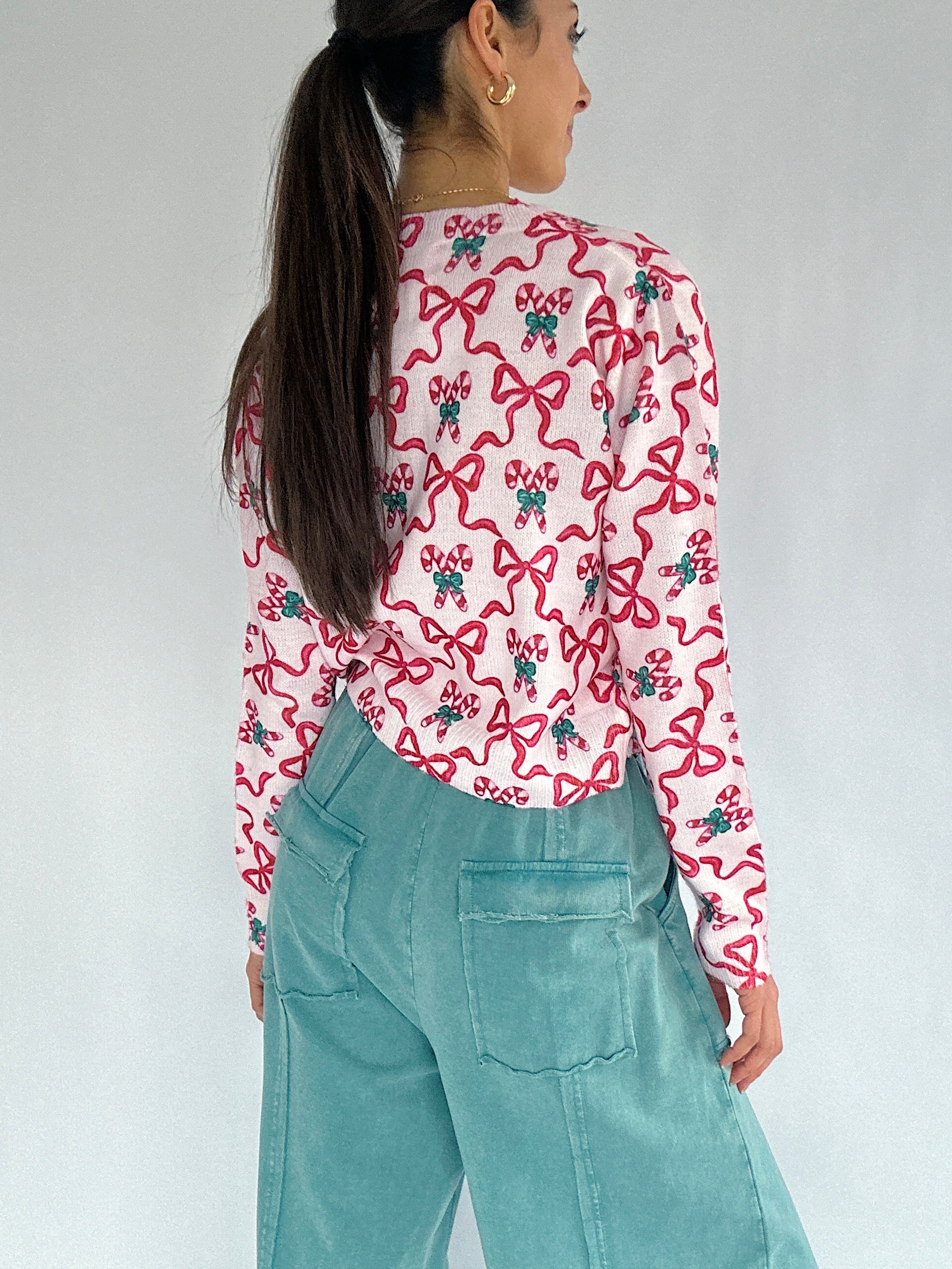 Person wearing a cardigan with red bow and candy cane pattern and teal pants against a plain background