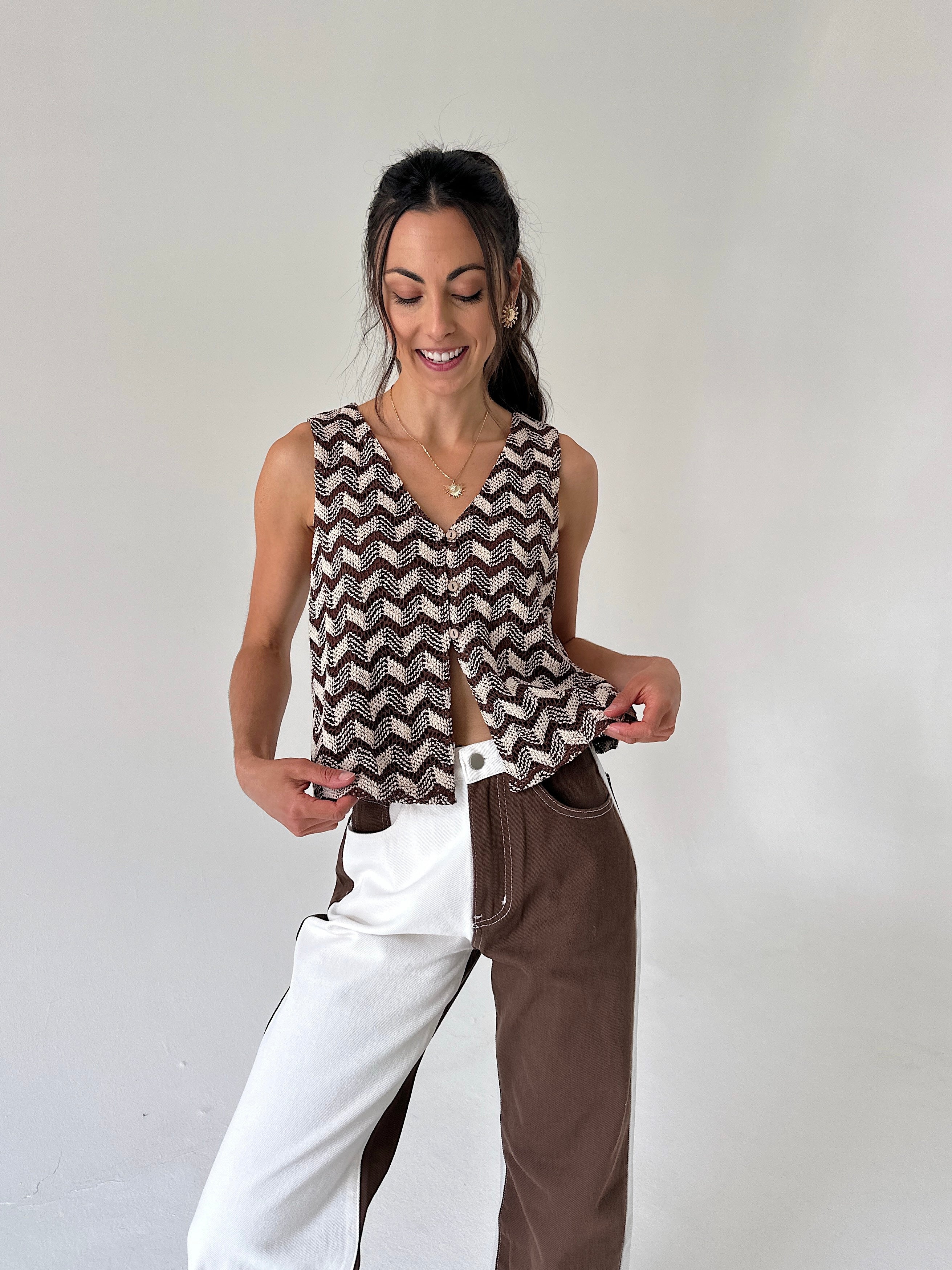 A woman modeling a sleeveless V-neck top with a brown and beige chevron pattern, paired with a split-color pants outfit, against a plain light gray background.