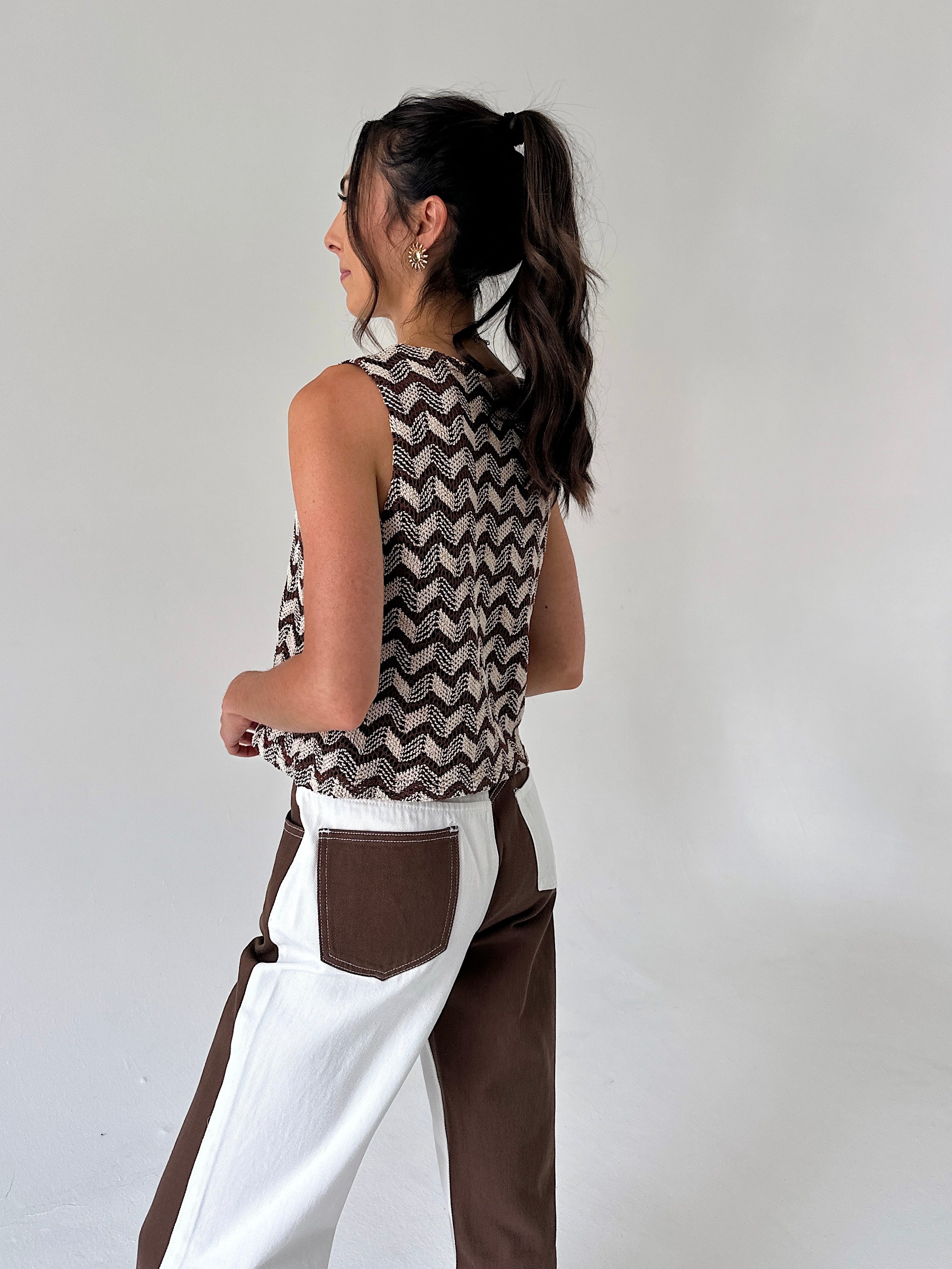 A woman shown from the back wearing a sleeveless knit top with a brown and white chevron pattern, paired with white and brown color-blocked pants, against a plain light gray background.