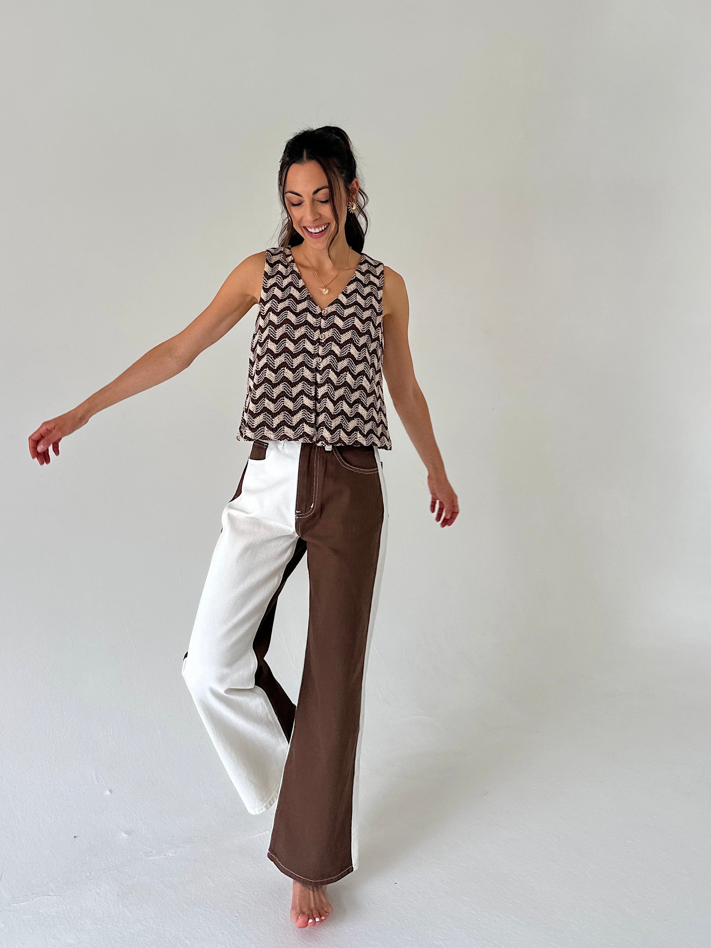 A woman modeling a sleeveless V-neck top with a brown and beige chevron pattern, paired with wide-leg pants split down the middle into white and brown sections, against a plain light gray studio background.
