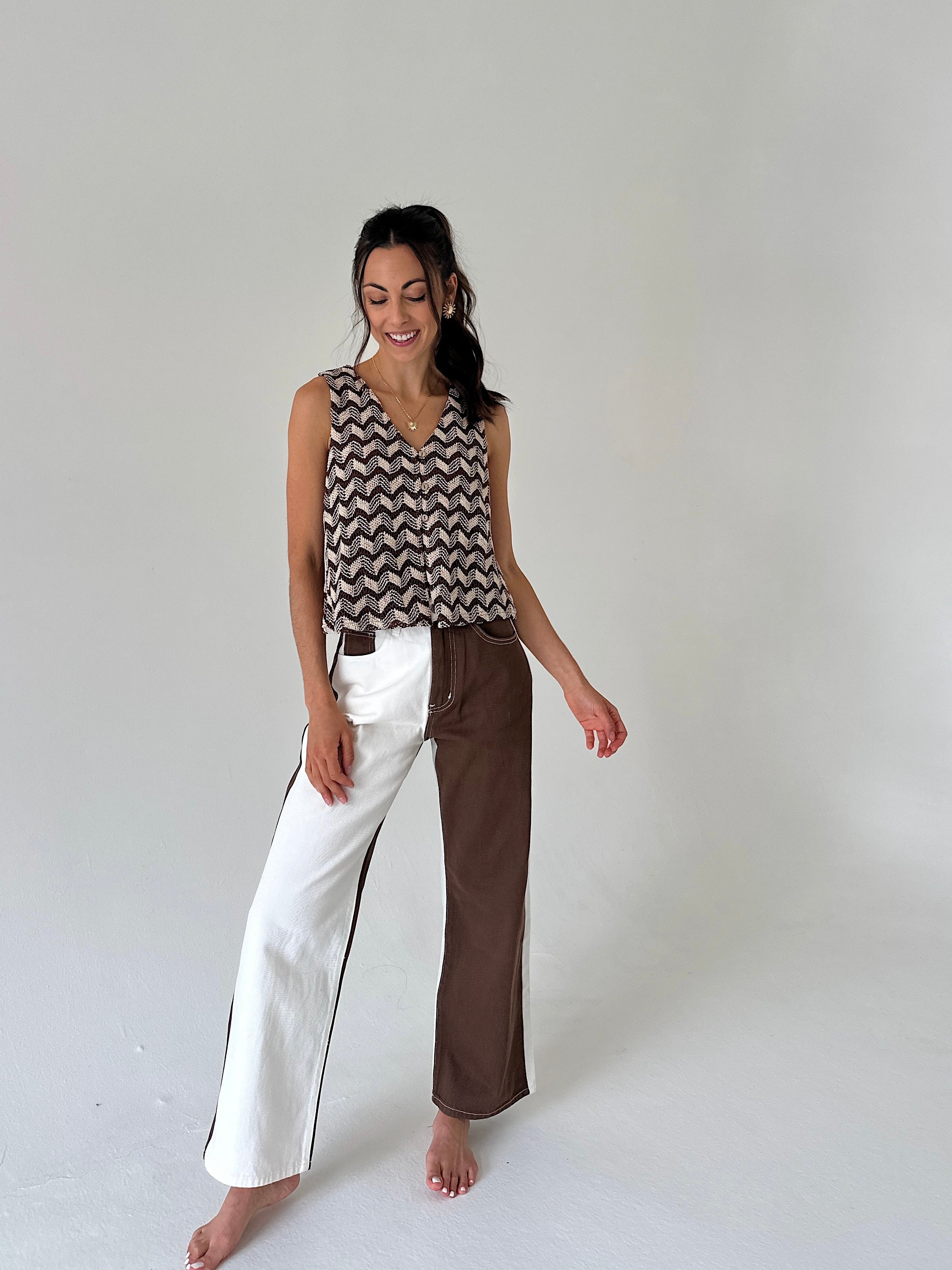 A woman stands barefoot against a plain light gray studio background, wearing a sleeveless top with a brown and beige chevron pattern and wide-leg pants split down the middle with one leg in white and the other in brown.