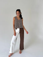 A woman stands barefoot against a plain light gray studio background, wearing a sleeveless top with a brown and beige chevron pattern and wide-leg pants split down the middle with one leg in white and the other in brown.