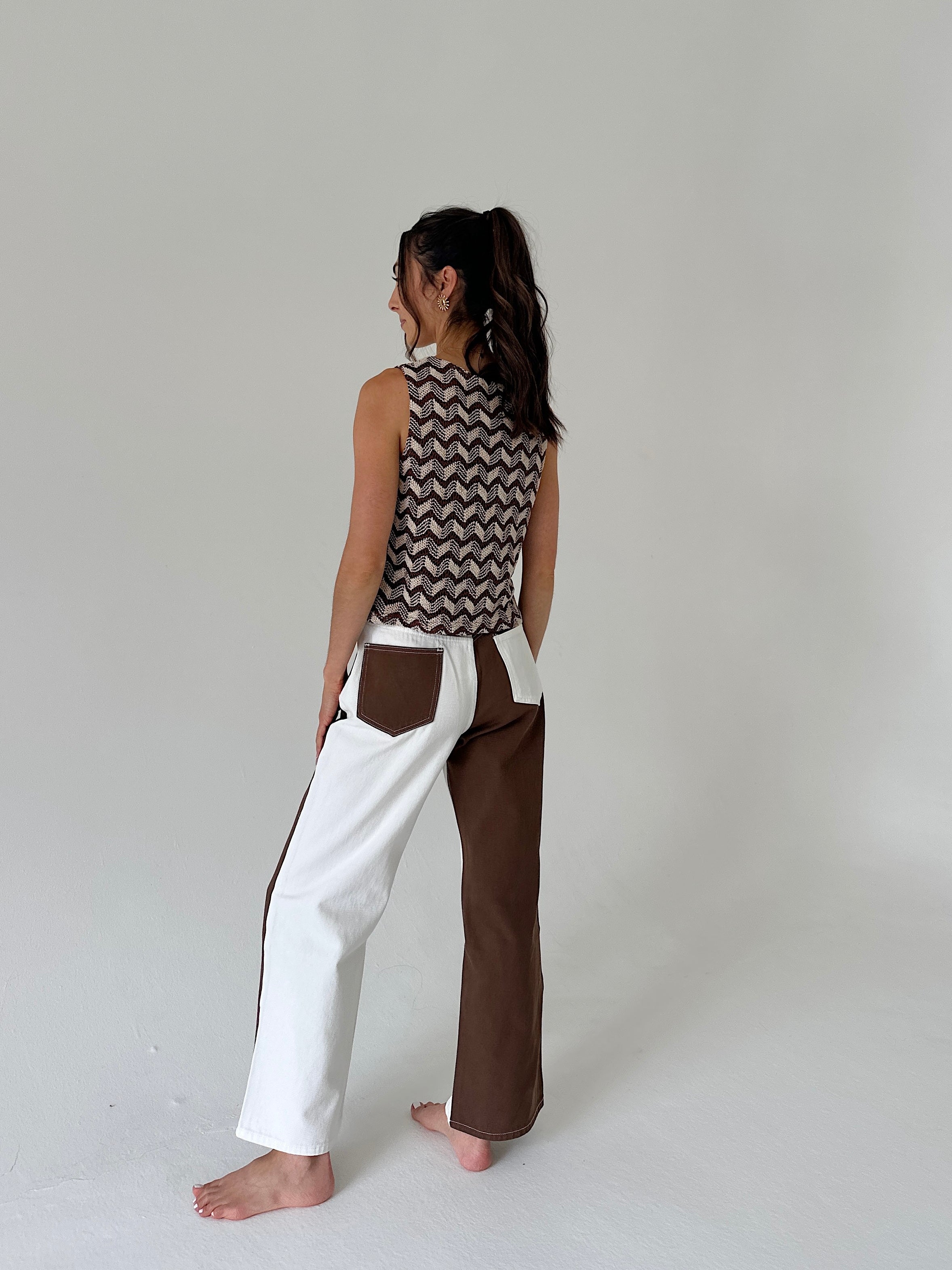 A woman stands barefoot in a studio wearing a sleeveless top with a brown and beige chevron pattern and wide-leg pants with a color-blocked design in white and brown.