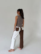 A woman stands barefoot in a studio wearing a sleeveless top with a brown and beige chevron pattern and wide-leg pants with a color-blocked design in white and brown.