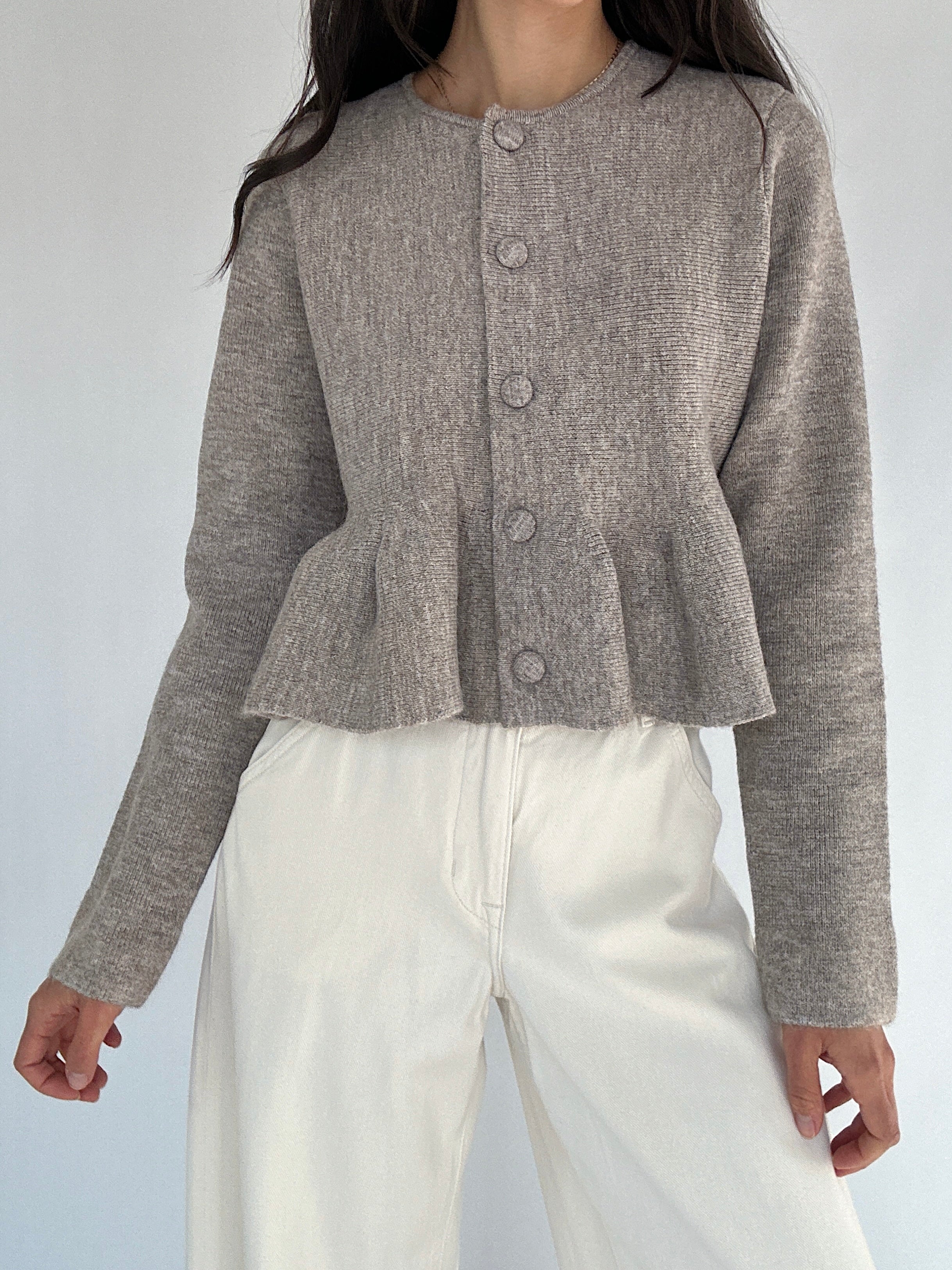 Person wearing a brown peplum cardigan top over white pants on a light gray background