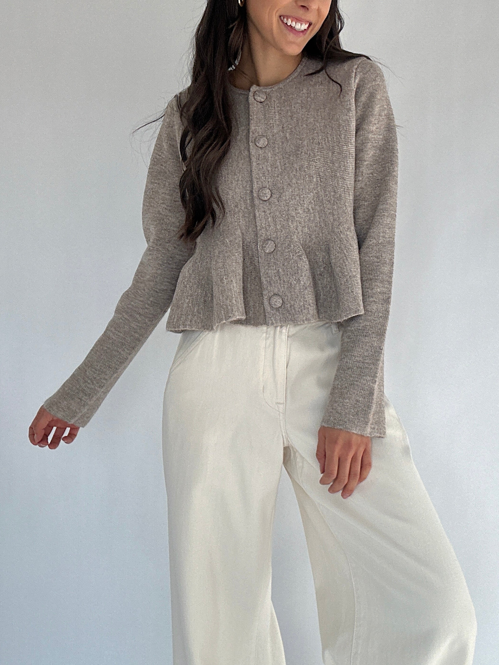 Woman wearing a brown peplum cardigan and white pants against a plain background