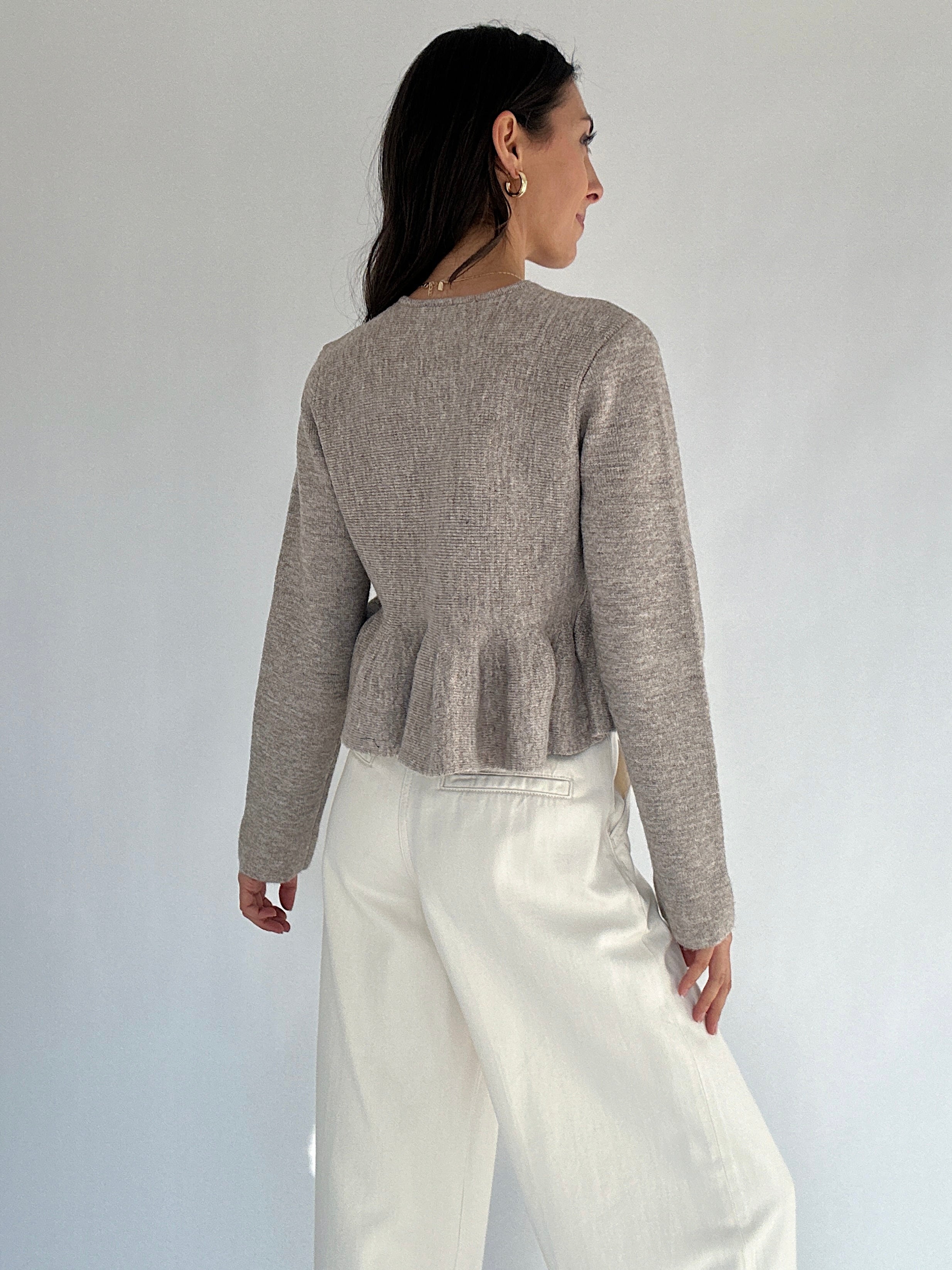 Woman wearing a brown peplum sweater and white pants against a plain background from back