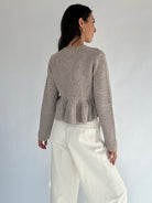 Woman wearing a brown peplum sweater and white pants against a plain background from back