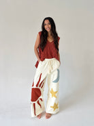 Woman wearing a rust-colored top and white pants with sun, moon and star designs on a plain background
