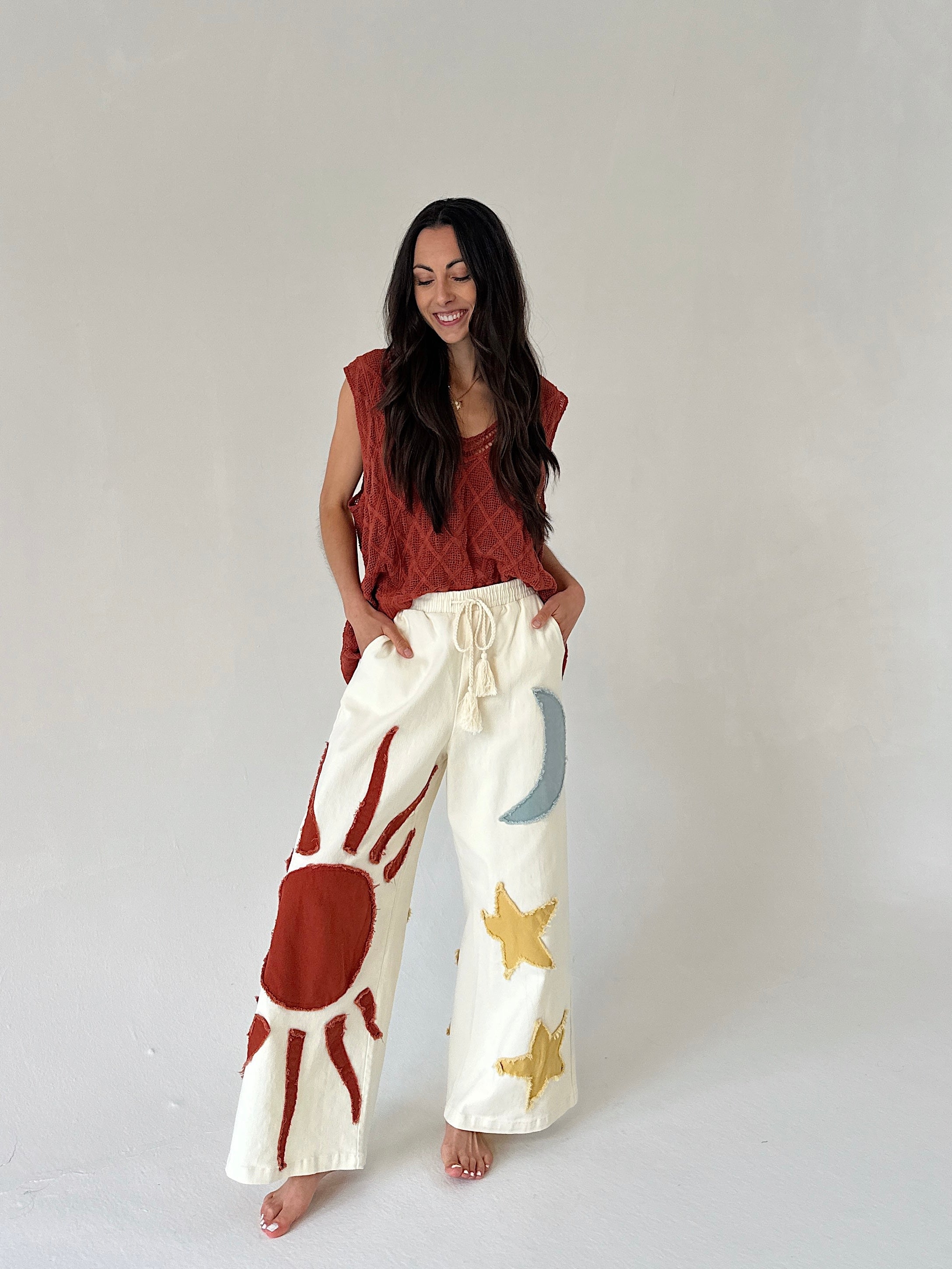 Woman wearing a rust-colored sleeveless top and white pants with sun and star patterns on a plain background