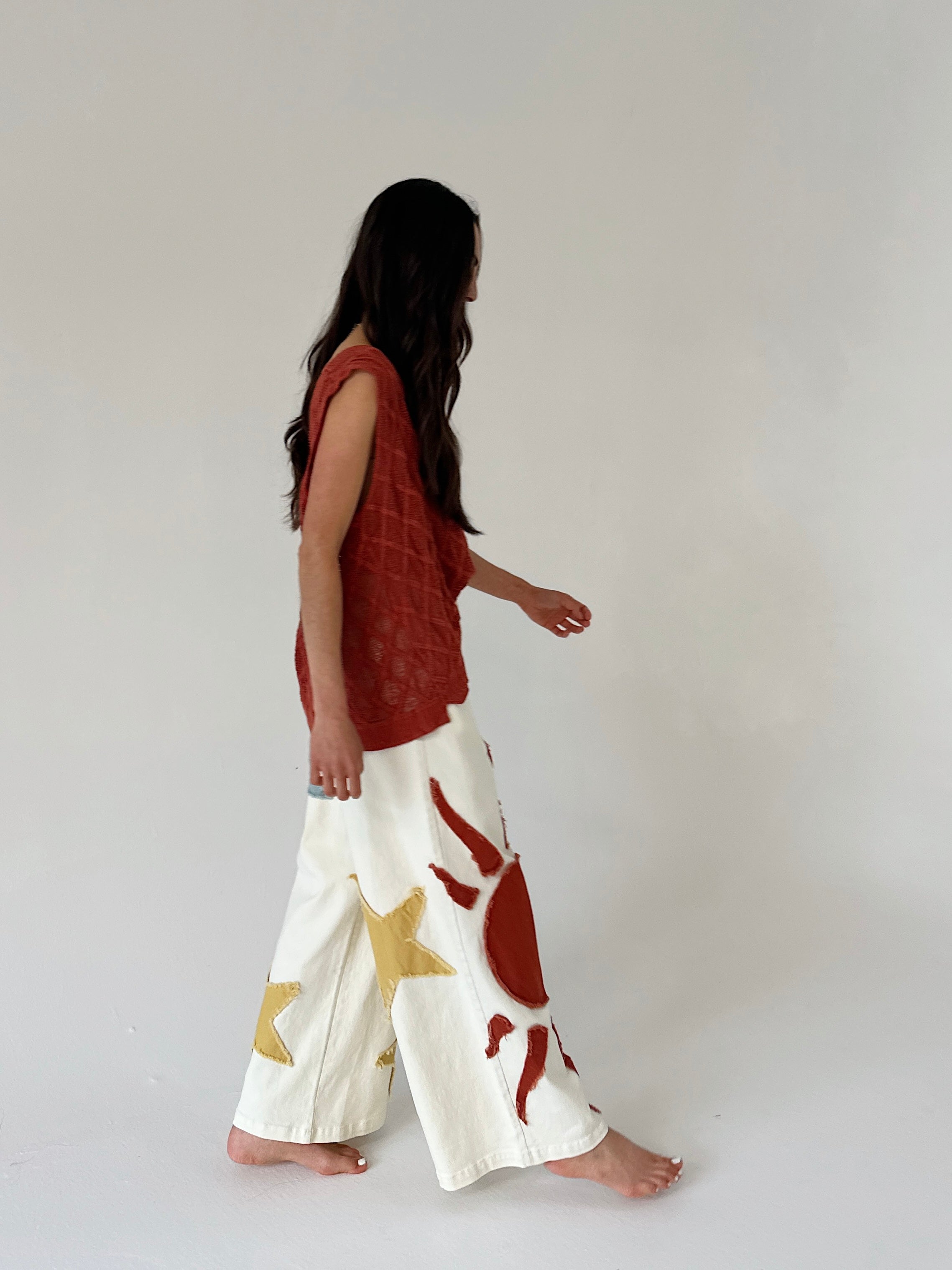 Woman wearing a rust-colored top and white pants with red and gold patterns on a plain background