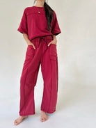 A person wearing a matching two-piece lounge set in deep burgundy, featuring an oversized short-sleeve top and wide-leg drawstring pants with side pockets, standing against a plain off-white background.