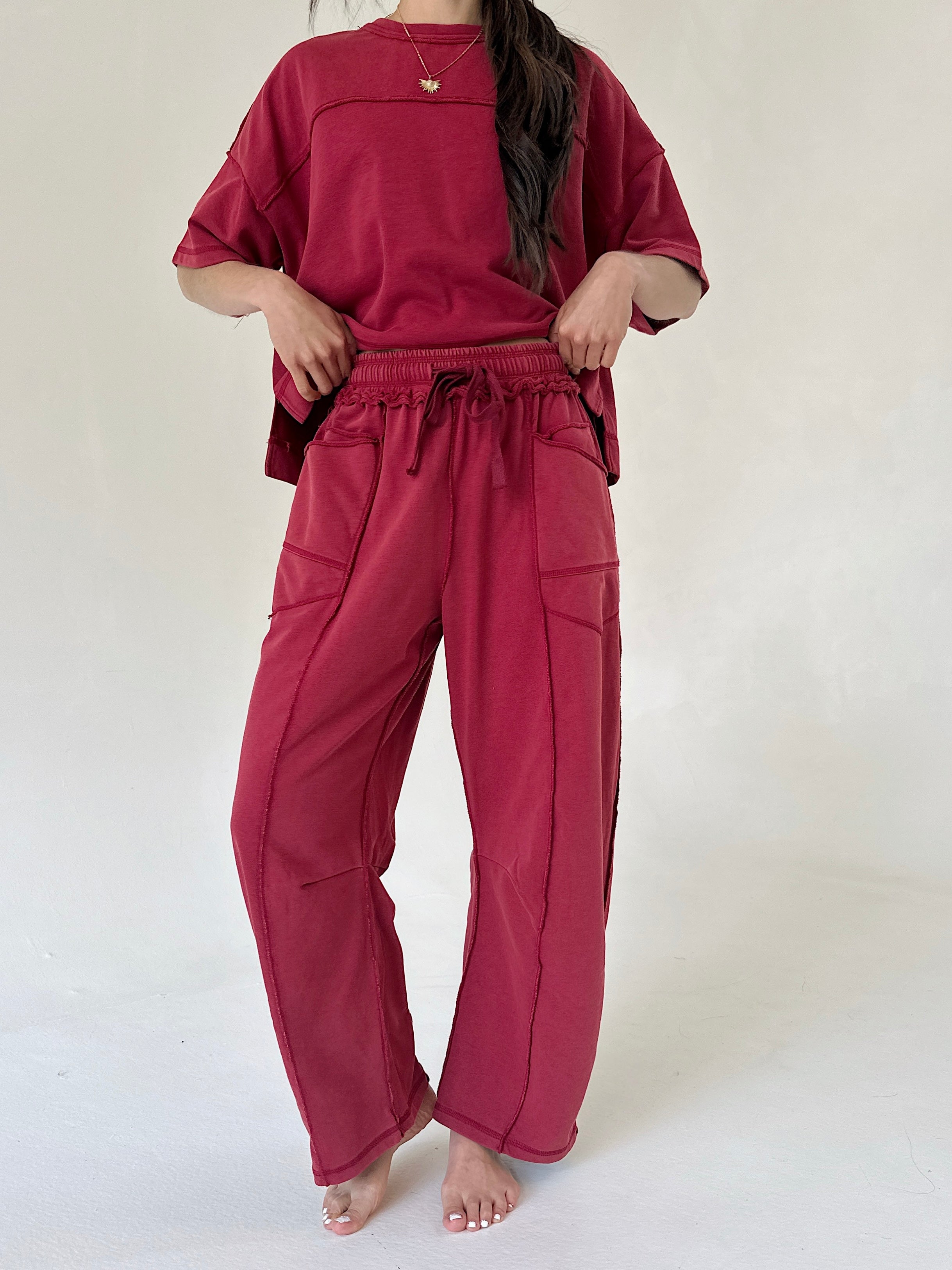 A woman modeling a matching two-piece lounge set in a deep berry red color, featuring an oversized short-sleeve top and wide-leg drawstring pants, standing against a plain off-white background.
