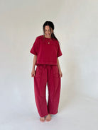 A woman modeling a matching two-piece lounge set in a deep red color, consisting of a short-sleeve cropped top and wide-leg pants with front patch pockets, standing against a plain off-white studio background.