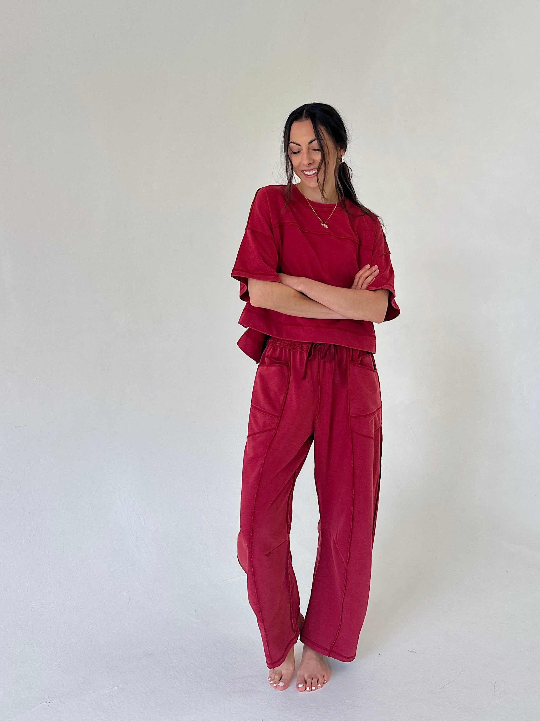 A woman modeling a matching two-piece set in a deep burgundy red, consisting of a short-sleeve top and wide-leg pants, standing against a plain off-white studio background.