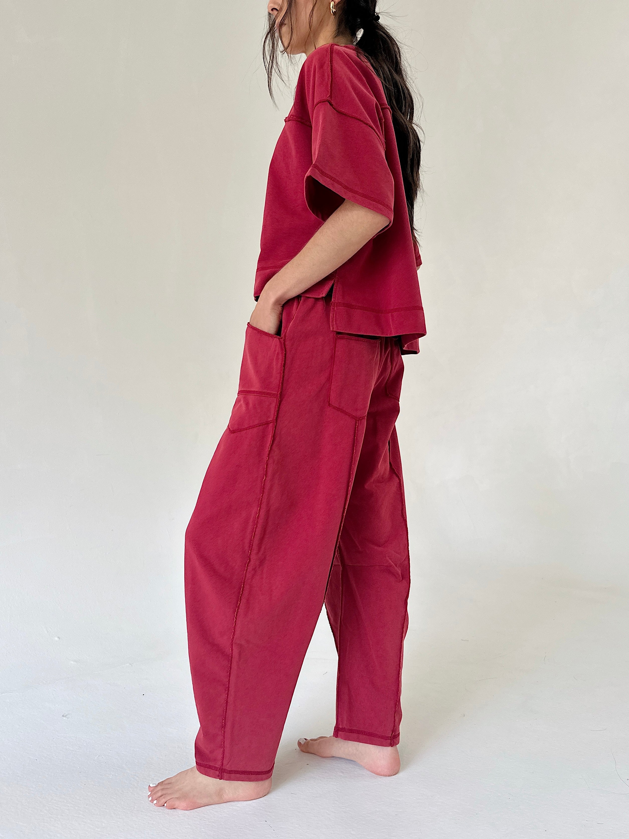 A woman wearing a matching two-piece lounge set in deep magenta, featuring a short-sleeve top and wide-leg pants with side pockets, standing barefoot against a plain off-white background.