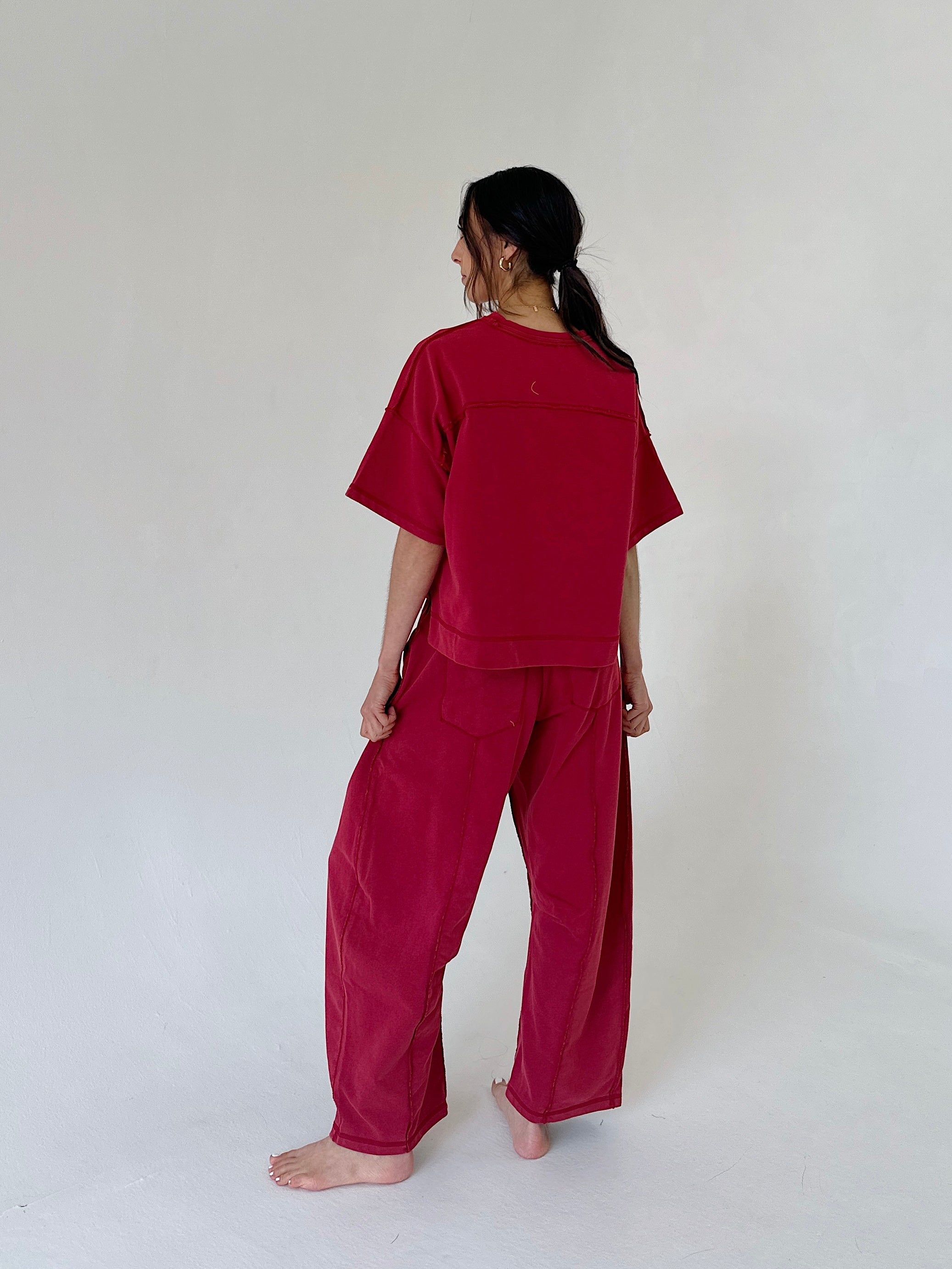 A woman stands barefoot against a plain off-white studio background, viewed from the back, wearing a loose-fitting red short-sleeve top and matching wide-leg pants.