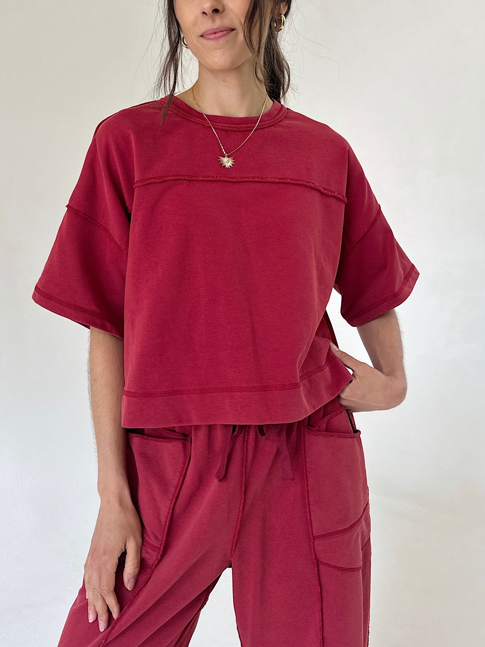 A woman wearing a cropped short-sleeve t-shirt and matching drawstring pants in a washed deep red or burgundy color, photographed against a plain off-white studio background.