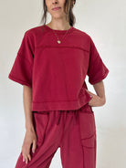A woman wearing a cropped short-sleeve t-shirt and matching drawstring pants in a washed deep red or burgundy color, photographed against a plain off-white studio background.