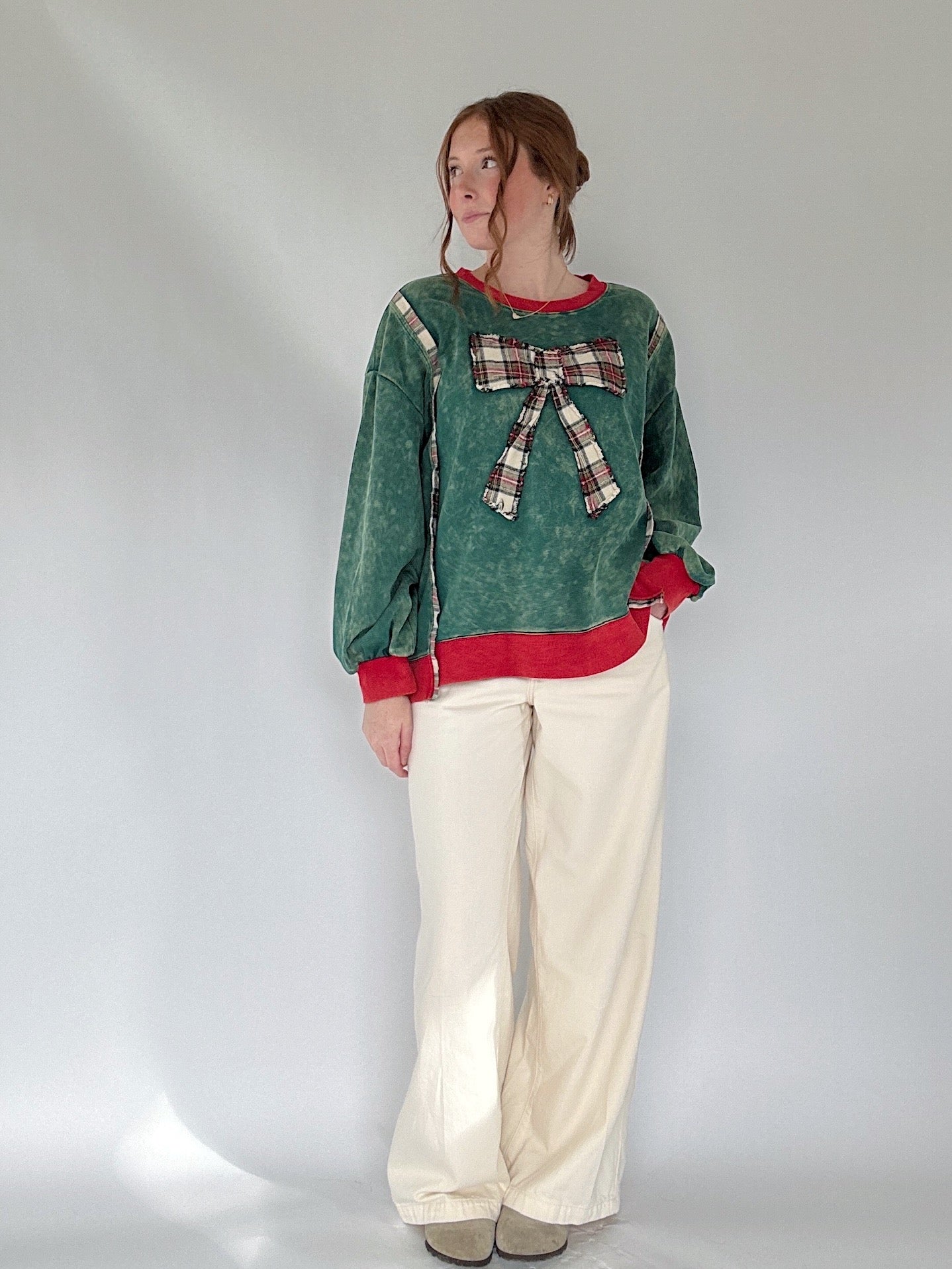 Person wearing a green pullover with a plaid bow and white pants on a plain background