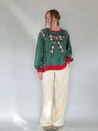 Person wearing a green pullover with a plaid bow and white pants on a plain background