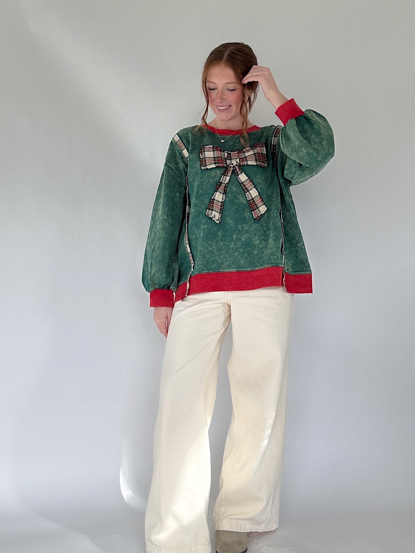 Person wearing a green pullover with a plaid bow and white pants on a plain background