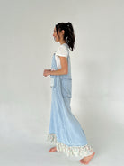 Woman wearing a light blue overalls with floral ruffled hem on a plain background from side