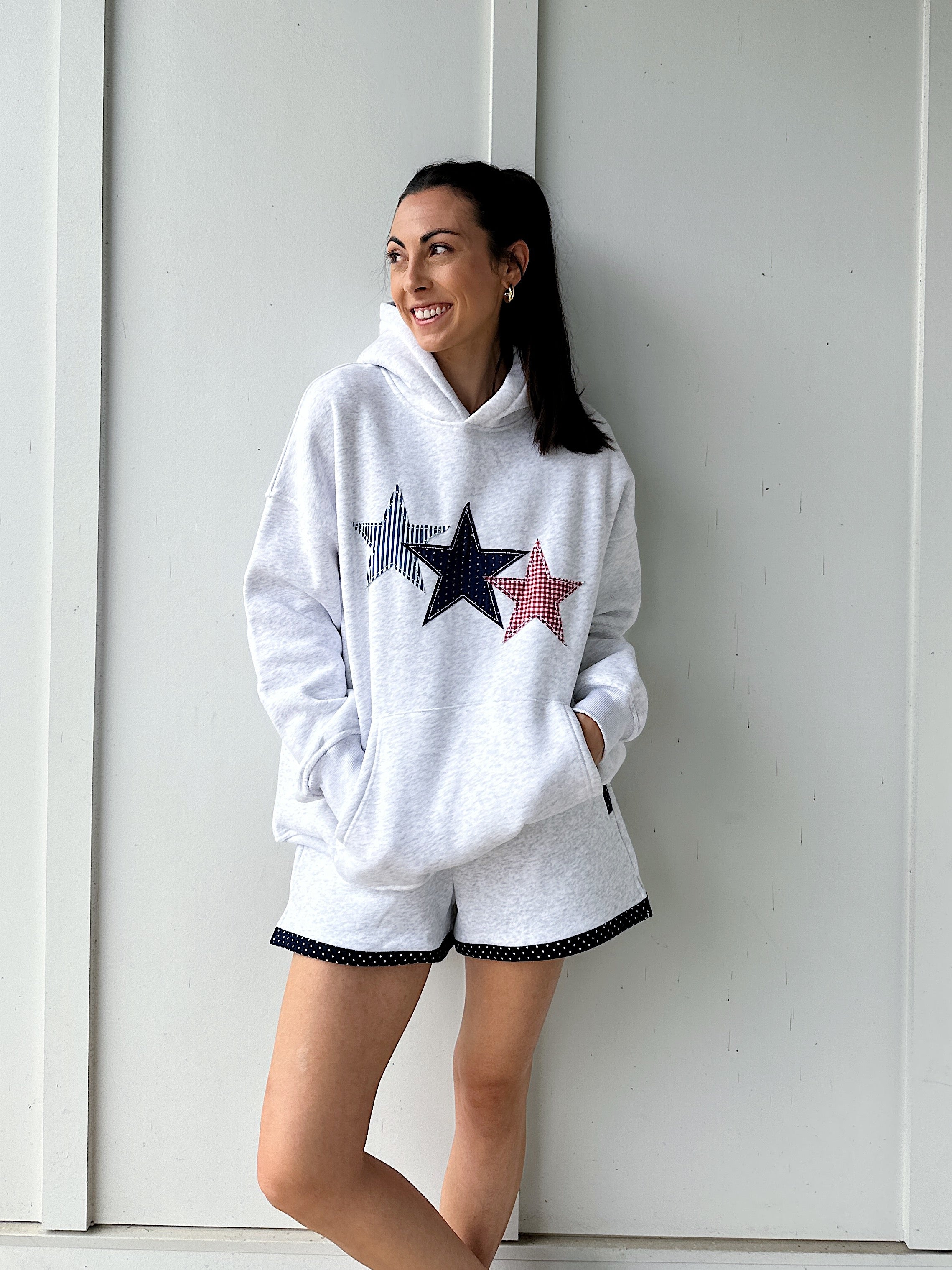 Woman wearing a grey hoodie with star designs and matching shorts against a light-colored wall.