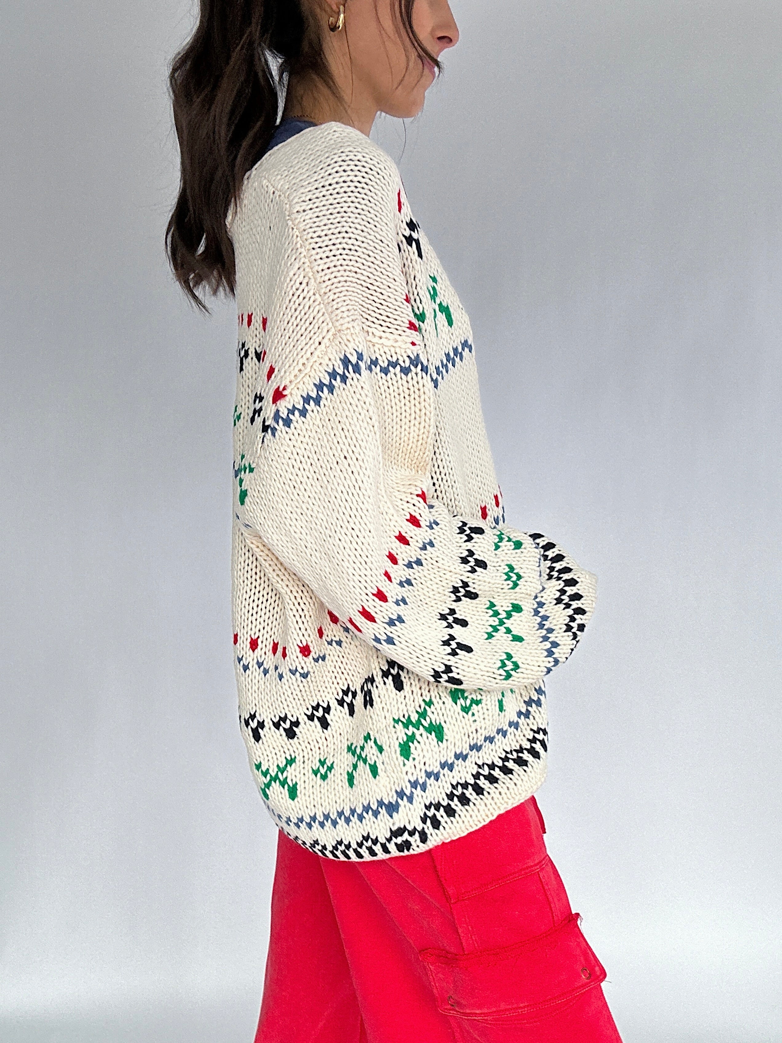 Person wearing a cream-colored knit sweater with colorful patterns and red pants on a plain background