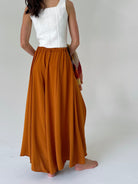 A woman shown from the back wearing a white sleeveless top and long, flowing rust-orange wide leg pants, standing barefoot against a plain light gray studio background.