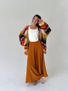 A woman stands barefoot against a light gray studio background, wearing a white sleeveless top, a long rust-orange maxi skirt, and a chunky multicolor striped cardigan with orange, yellow, red, green, and brown tones.
