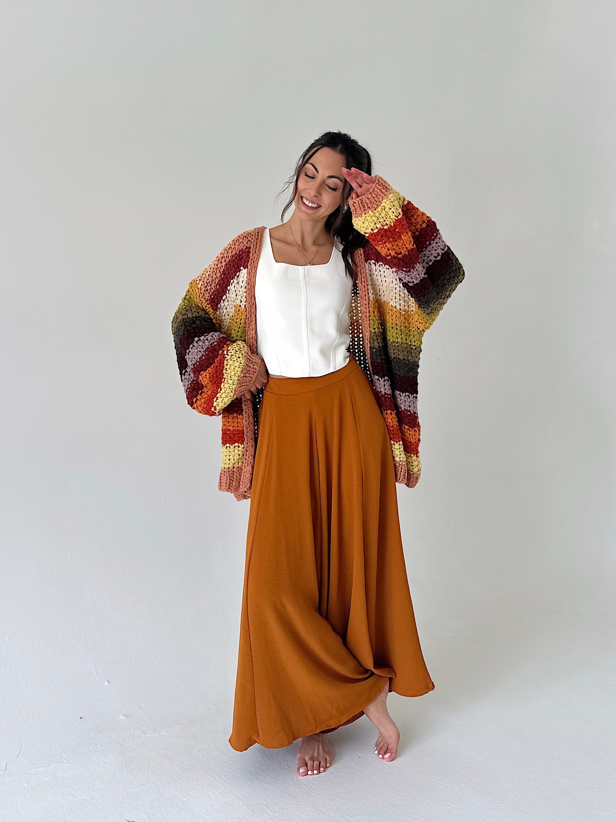 A woman stands barefoot against a light gray studio background, wearing a white sleeveless top, a long rust-orange maxi skirt, and a chunky multicolor striped cardigan with orange, yellow, red, green, and brown tones.