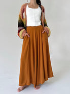 A woman modeling a long, flowing rust-orange wide leg pants with side pockets, paired with a white sleeveless top and a chunky multicolor knit cardigan with striped sleeves, standing against a light gray studio background.