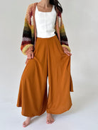 A woman modeling a pair of wide-leg rust-colored pants with a white top and a multicolor striped knit cardigan, standing against a plain light gray background.