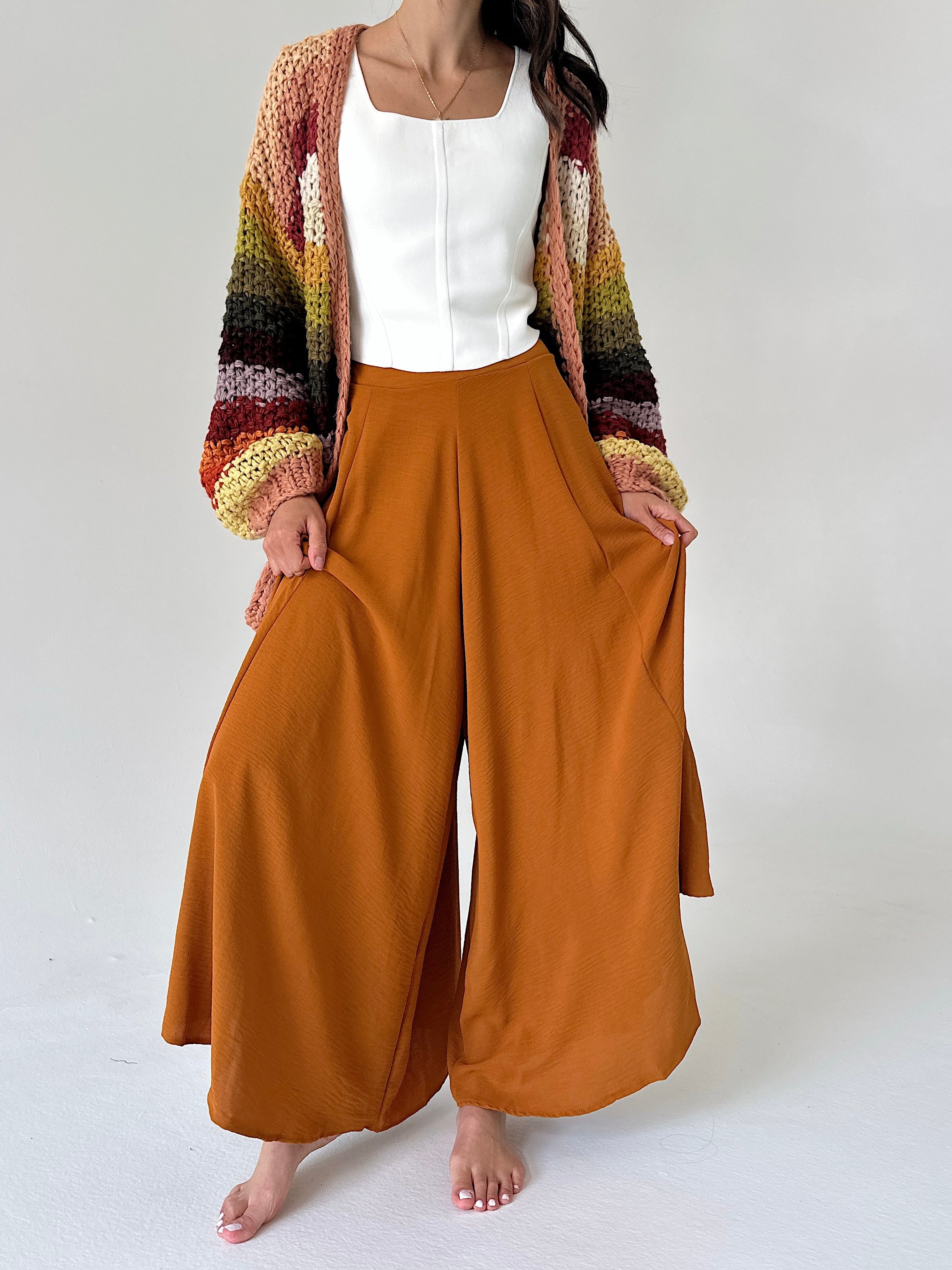 A woman modeling a pair of wide-leg rust-colored pants with a white top and a multicolor striped knit cardigan, standing against a plain light gray background.