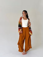 A woman stands barefoot against a plain light gray studio background, wearing a white sleeveless top, wide-leg rust-brown pants, and a chunky multicolor knit cardigan draped off her shoulders.