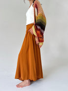 A model wearing a rust-orange maxi pant, a white top, and a chunky multicolor crochet cardigan draped over one shoulder, standing barefoot against a light gray studio background.