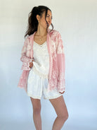 Woman wearing a pink lace button down over a white outfit on a plain background
