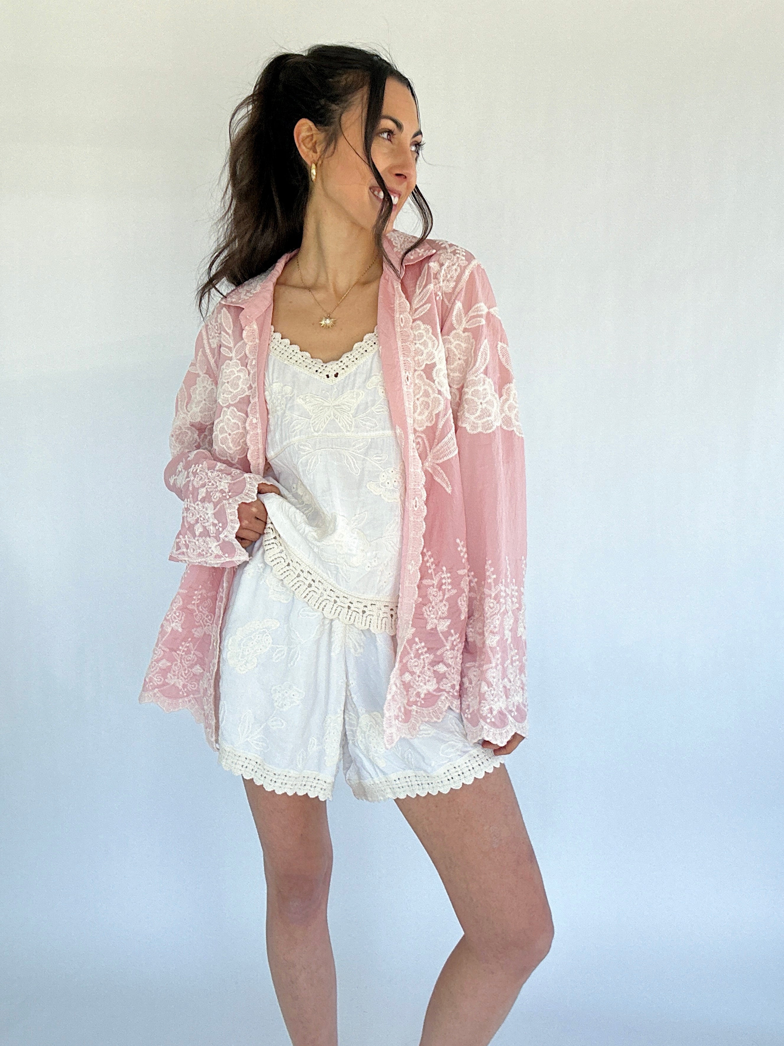 Woman wearing a pink lace button down over a white outfit on a plain background