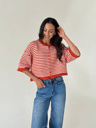 A woman modeling a cropped orange and white striped knit cardigan with blue jeans, standing against a plain off-white wall.