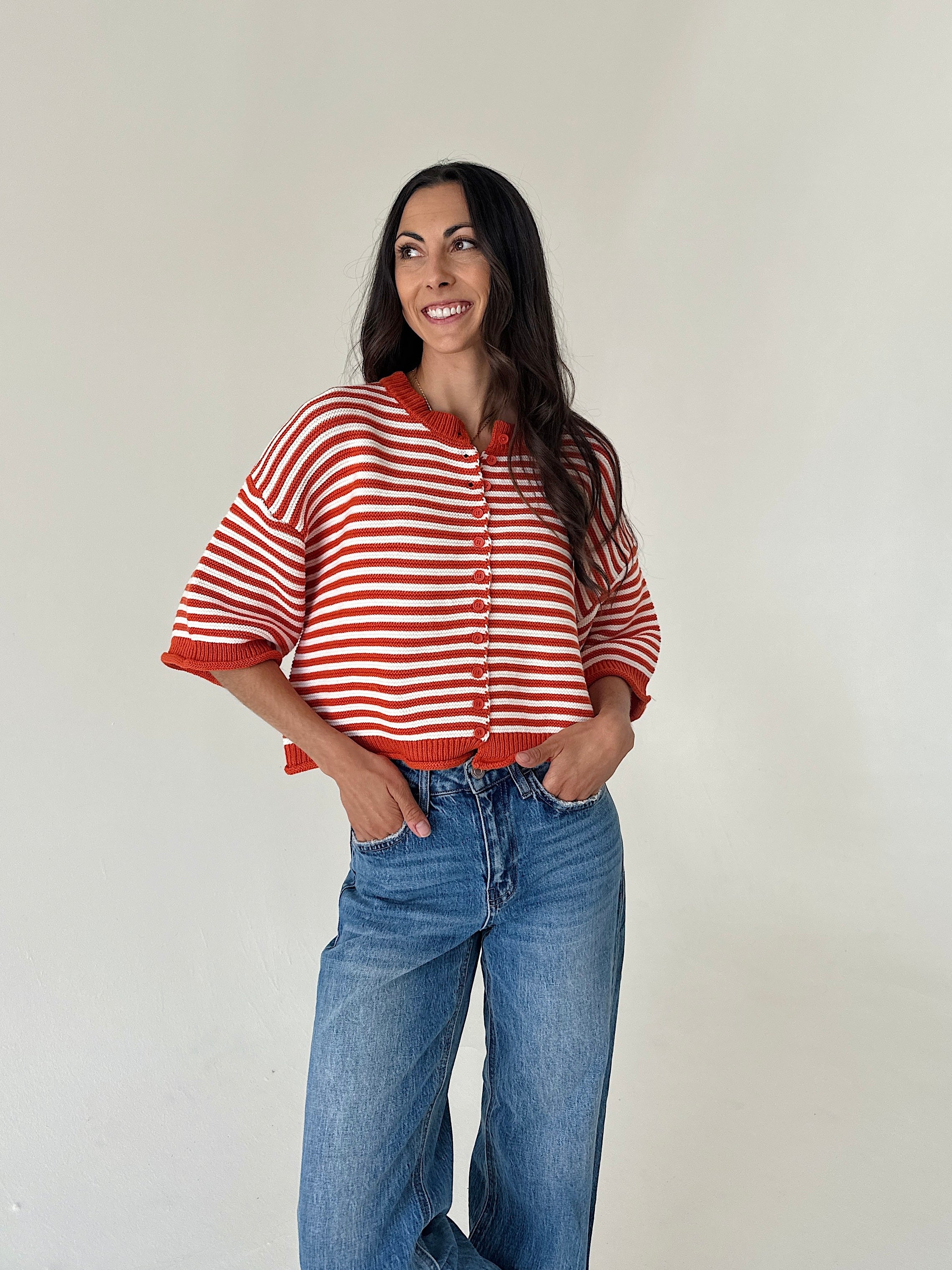 A woman modeling an orange and white striped knit cardigan with a drawstring neckline, paired with blue jeans, against a plain off-white wall.