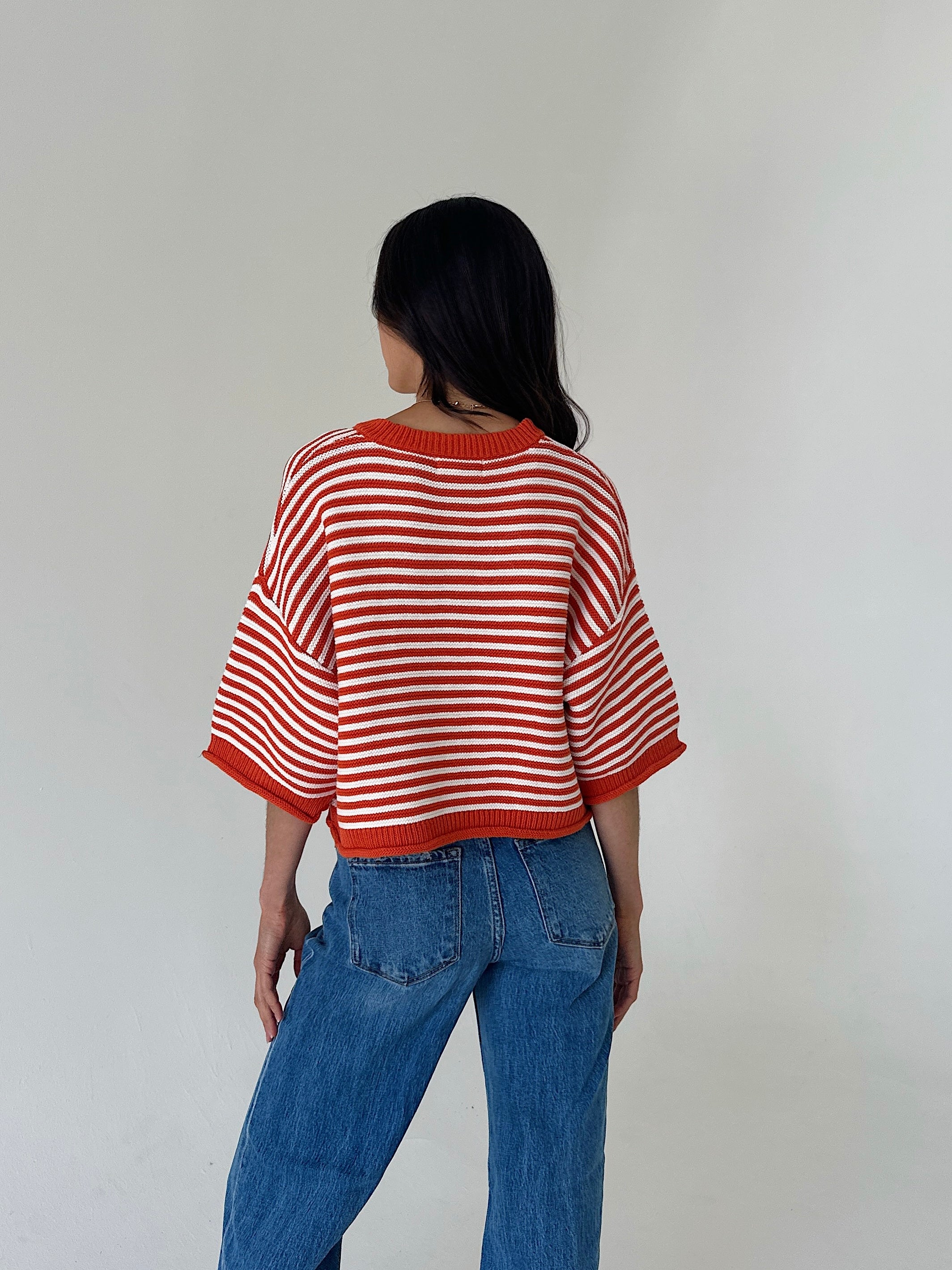 A woman viewed from behind wearing an orange and white striped short-sleeve knit top and blue jeans, standing against a plain light gray wall.