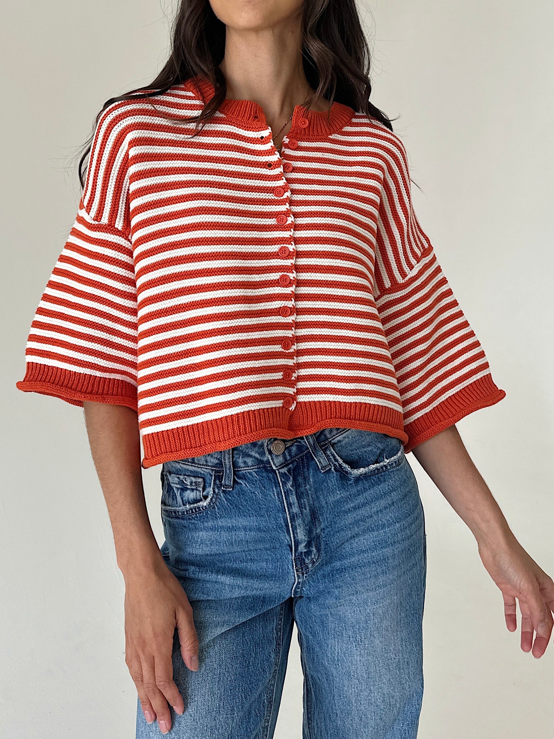 A woman wearing a cropped, short-sleeve knit cardigan with red and white horizontal stripes, paired with blue jeans, against a plain light background.