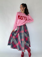 Woman wearing a pink 'Merry' sweater and plaid skirt on a white background