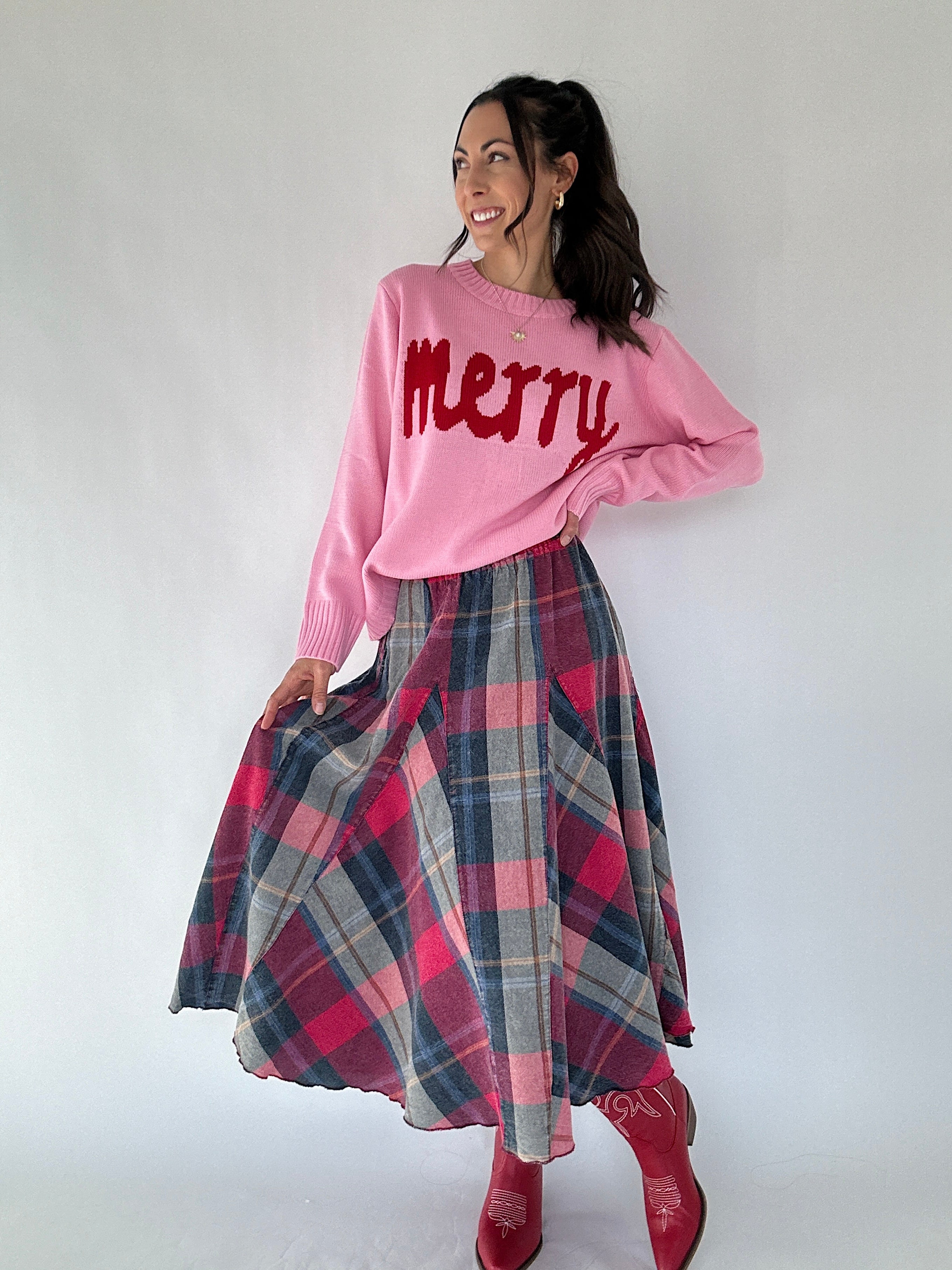 Woman wearing a pink 'Merry' sweater and plaid skirt on a white background