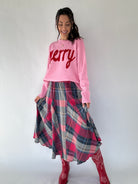 Woman wearing a pink sweater with 'Merry' text and a plaid skirt on a white background