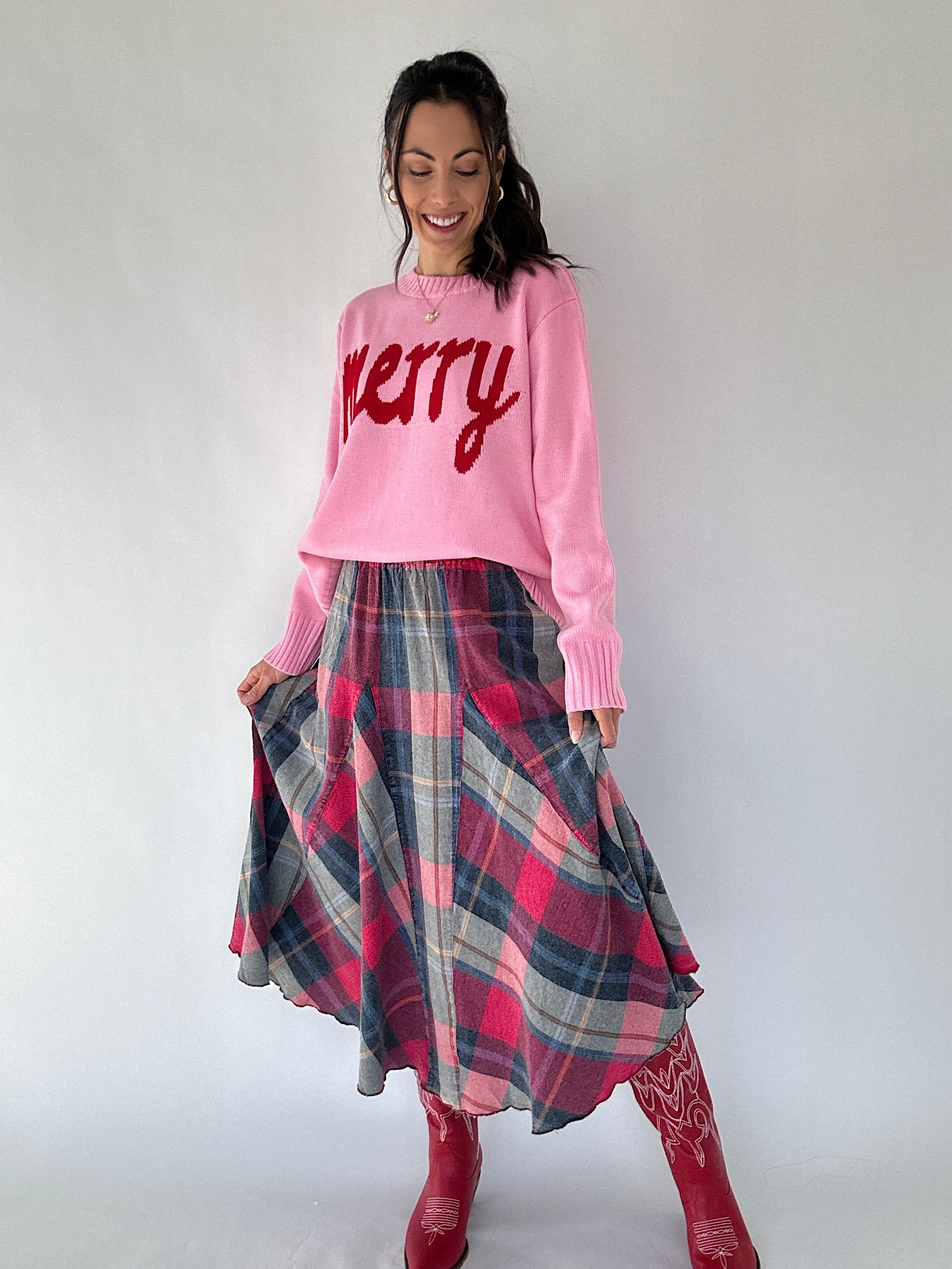 Woman wearing a pink sweater with 'Merry' text and a plaid skirt on a white background